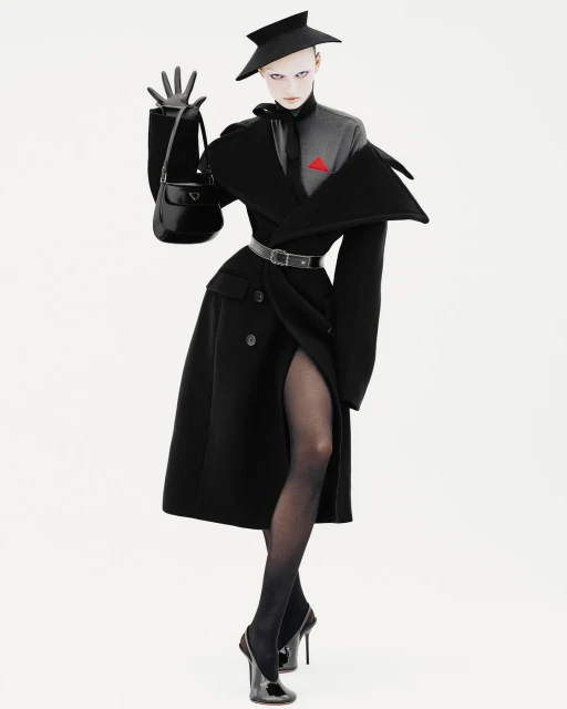 Fashion editorial shot of an adult female wearing a black coat, hat, gloves, and heels, posing with a black purse on a white background.
