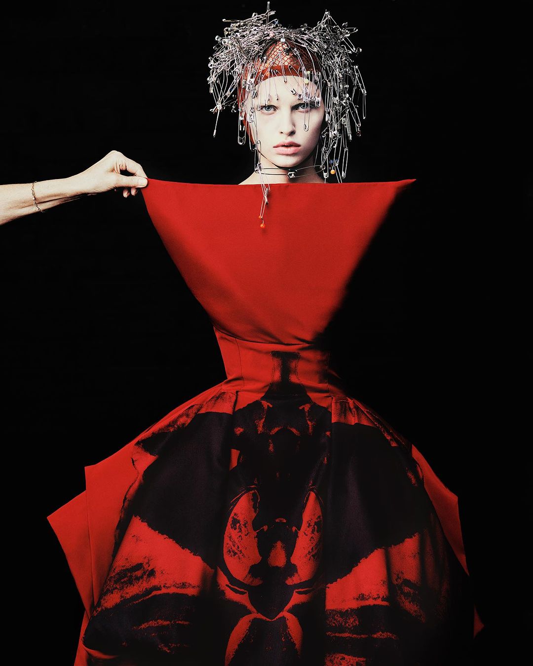 High-fashion portrait of a woman in a red dress and silver hairpin headpiece, partially revealed by a hand against a black backdrop.