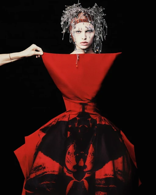 High-fashion portrait of a woman in a red dress and silver hairpin headpiece, partially revealed by a hand against a black backdrop.