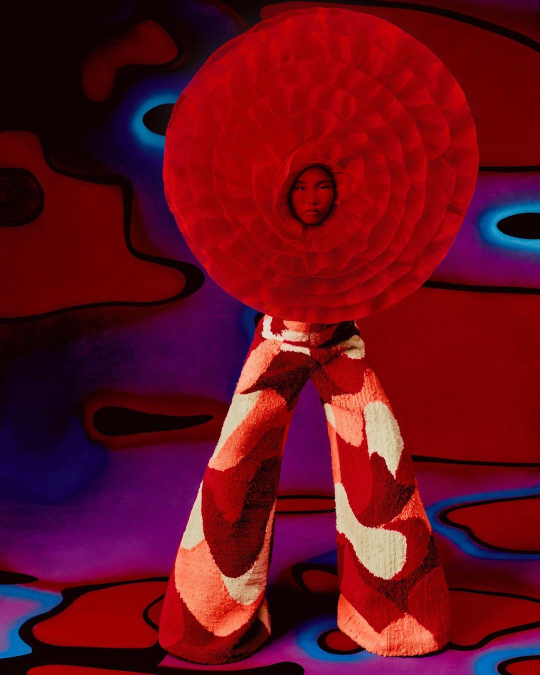 A person wearing a large round red flower-like headpiece and patterned pants stands against a surreal red and purple background.
