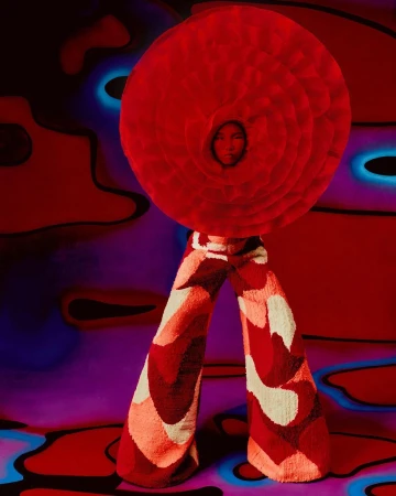 A person wearing a large round red flower-like headpiece and patterned pants stands against a surreal red and purple background.