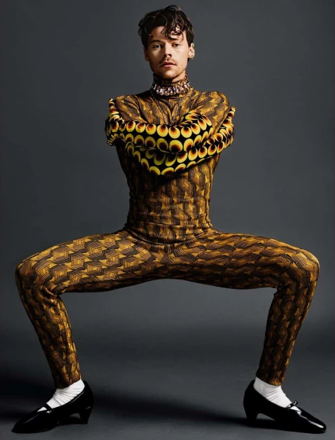 Studio portrait of a male model in a brown patterned bodysuit posing with spread legs and arms, black patent leather shoes, grey background.