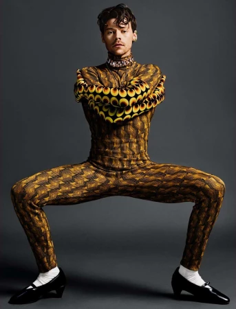 Studio portrait of a male model in a brown patterned bodysuit posing with spread legs and arms, black patent leather shoes, grey background.