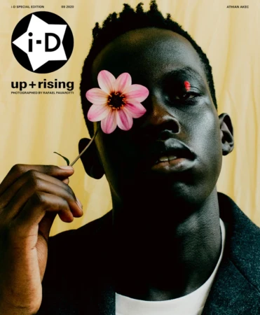 Portrait of a Black man holding a pink flower over his right eye against a warm textured yellow background, photographed by Rafael Pavarotti.