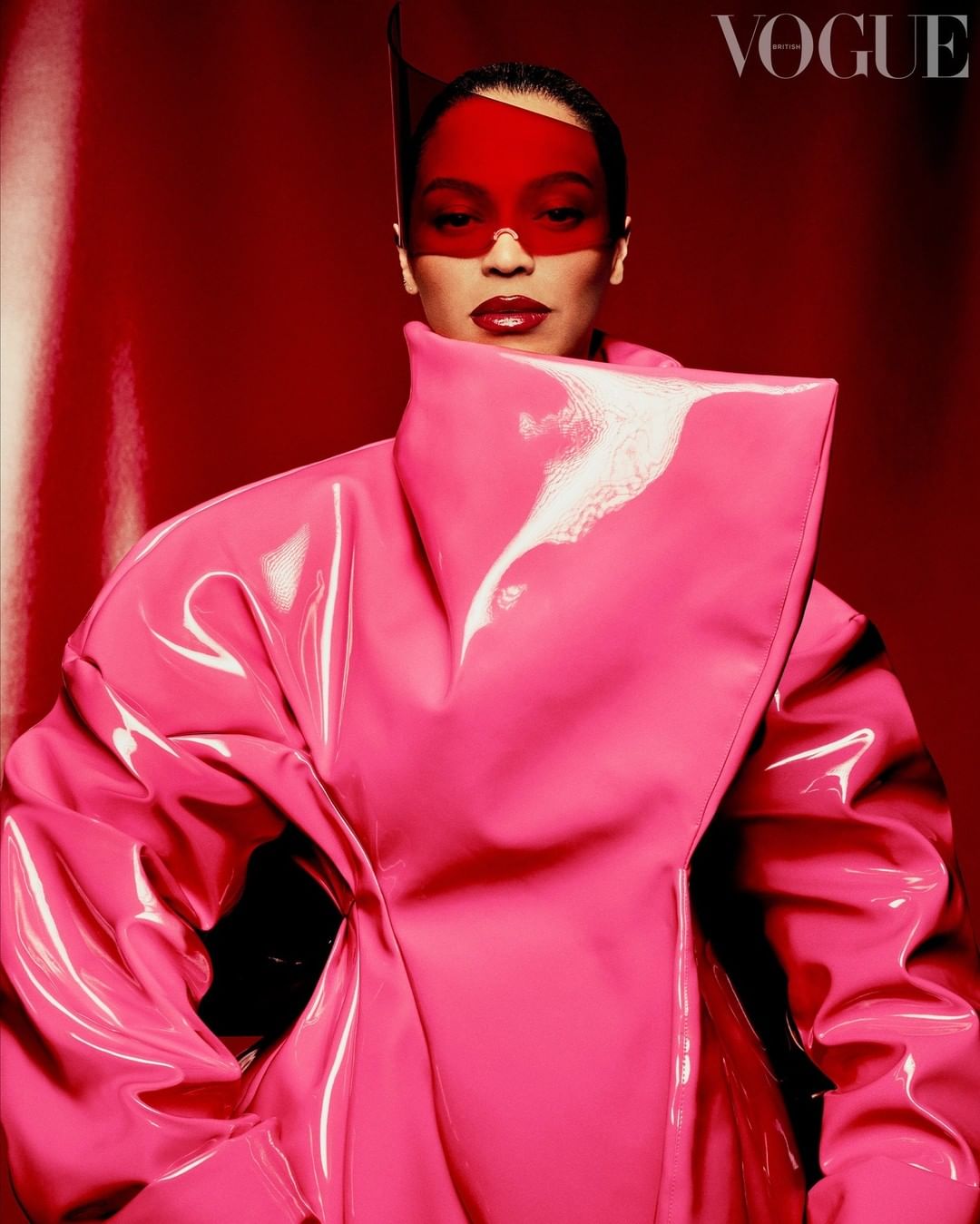 A woman with a visor and pink latex coat poses against a red background, as seen in a studio shot for a Vogue fashion editorial.