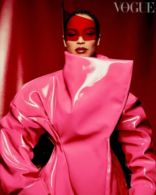 A woman with a visor and pink latex coat poses against a red background, as seen in a studio shot for a Vogue fashion editorial.