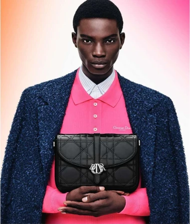 Dark-skinned male model poses in pink polo and textured blue coat holding black Dior handbag against gradient pink-orange background.