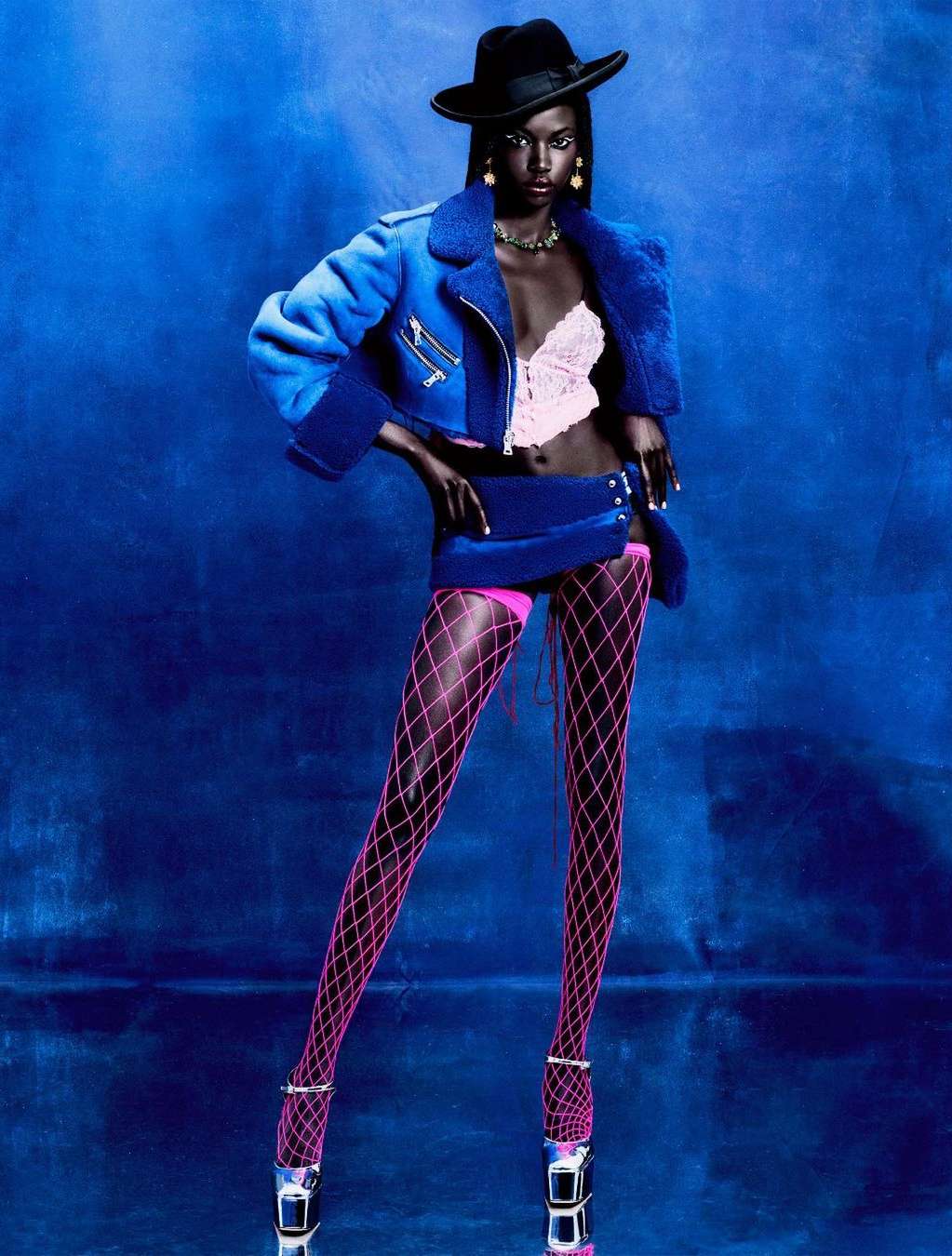 Full-length studio shot of a Black woman in a blue jacket and skirt, pink hosiery, and high heels against a blue background.