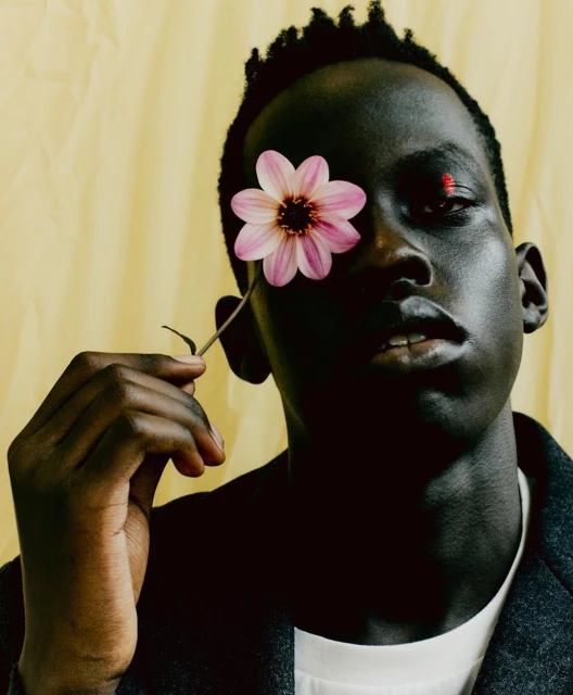 Close-up portrait of a dark-skinned Black male model holding a pink flower in front of one eye against a yellow backdrop.