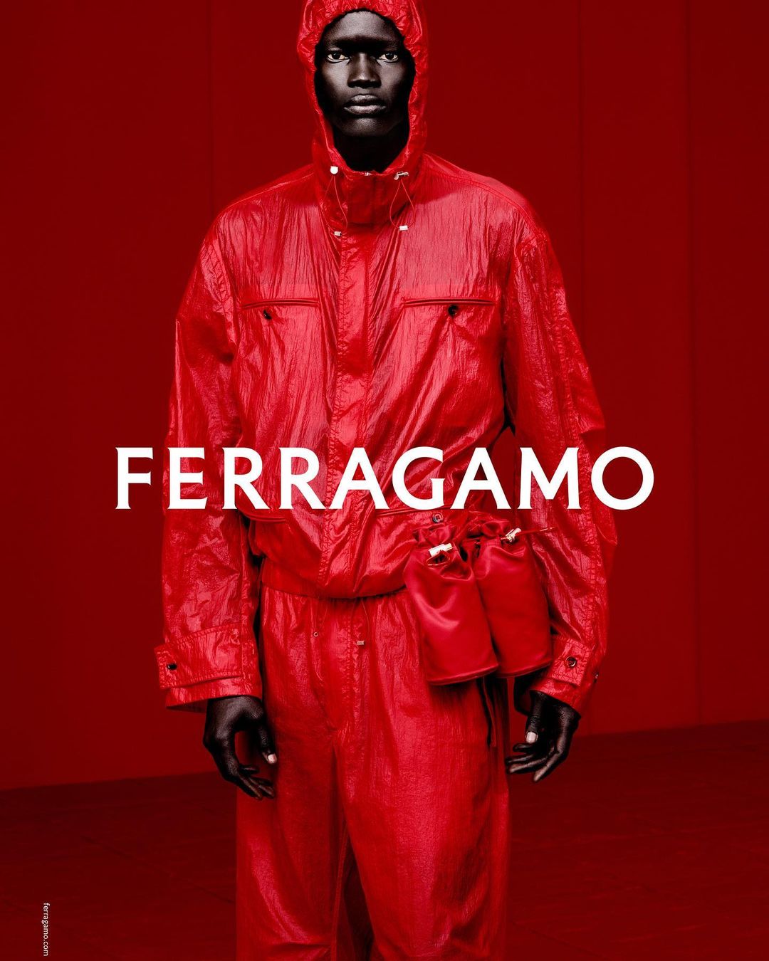 Fashion shot of a Black male model wearing a red hooded jacket and pants from Ferragamo against a red background, logo in white.