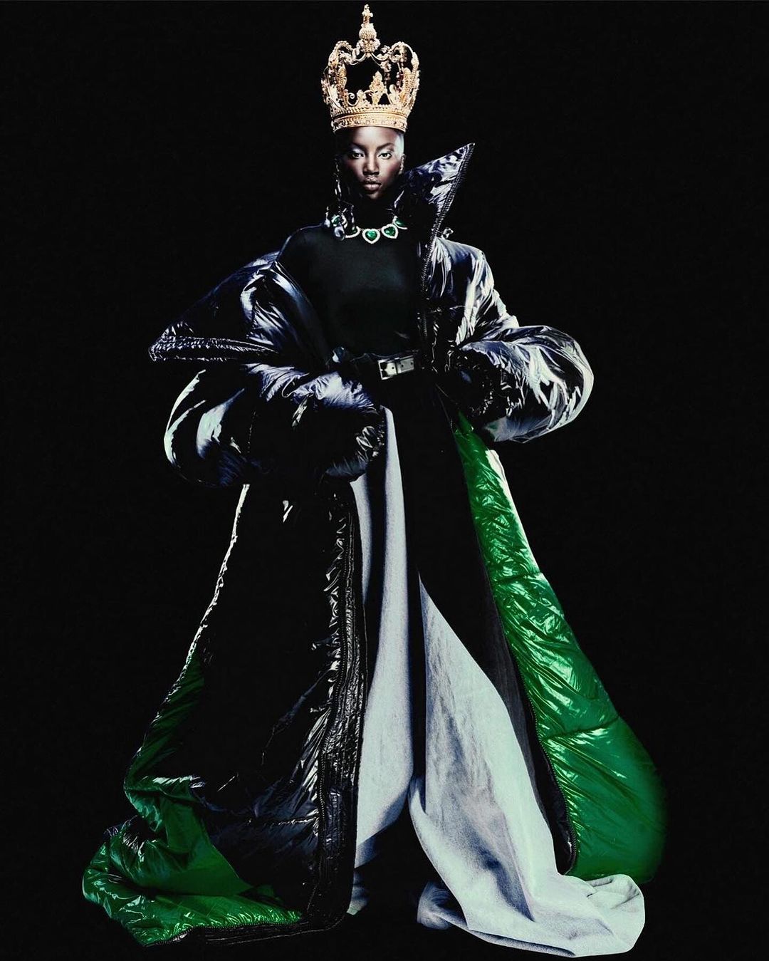 Fashionable Black woman in a stylish puffer coat and gold crown poses against a dark background, exuding modern regal elegance.