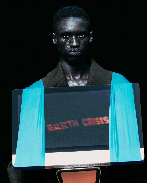 Fashion model presenting 'Earth Crisis' message on monitor, adorned with light blue ribbons, during a fashion show on a dark stage.