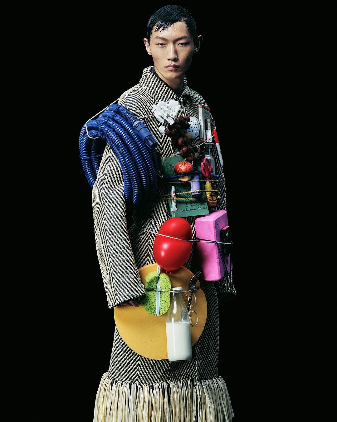 A person in a patterned coat poses in a studio against a black backdrop, with various objects attached to the clothing.