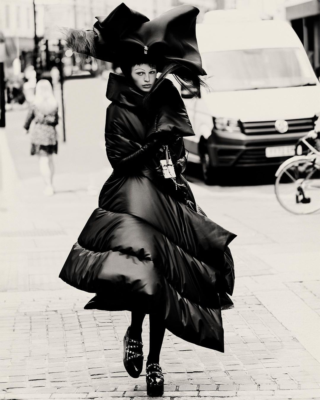 A person in a large black puffer coat and oversized hat walks on a cobblestone street with a white van in the background.