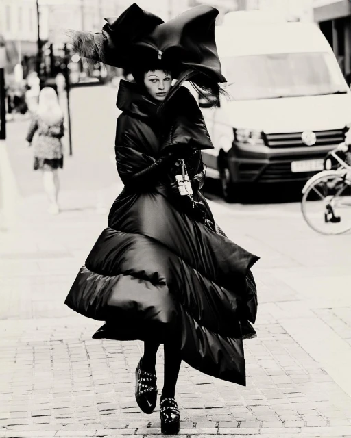 A person in a large black puffer coat and oversized hat walks on a cobblestone street with a white van in the background.