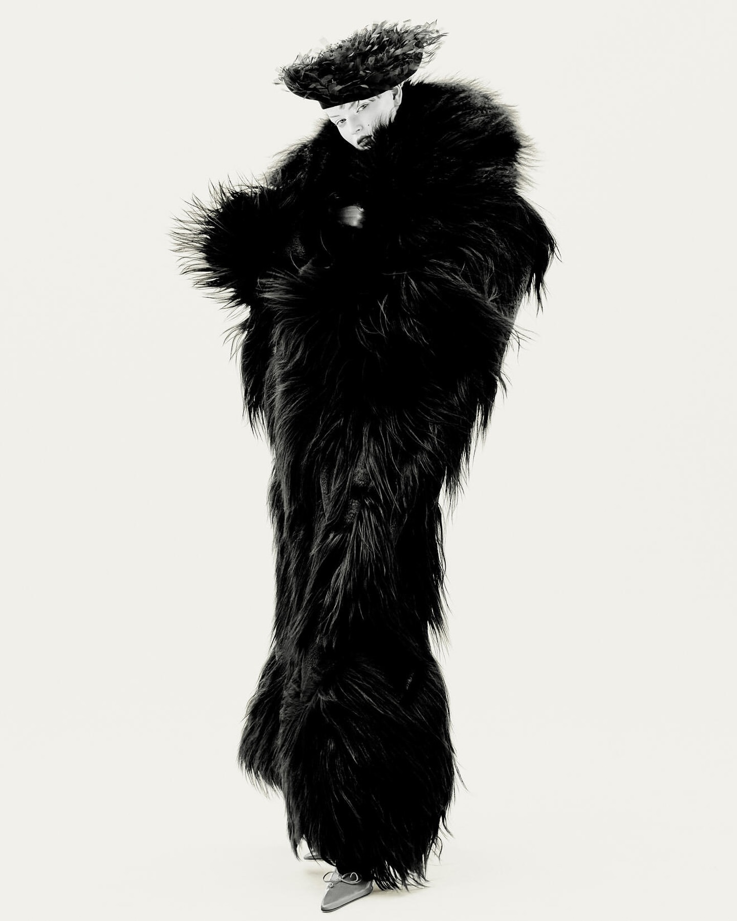 Full-length shot of a person wearing a furry black floor-length coat and matching hat, posing in a white studio setting.
