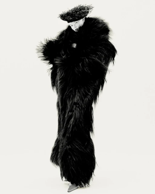 Full-length shot of a person wearing a furry black floor-length coat and matching hat, posing in a white studio setting.