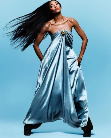 Fashion shot of Naomi Campbell in a draped blue gown against a blue backdrop, hands in pockets, with long black hair flowing.