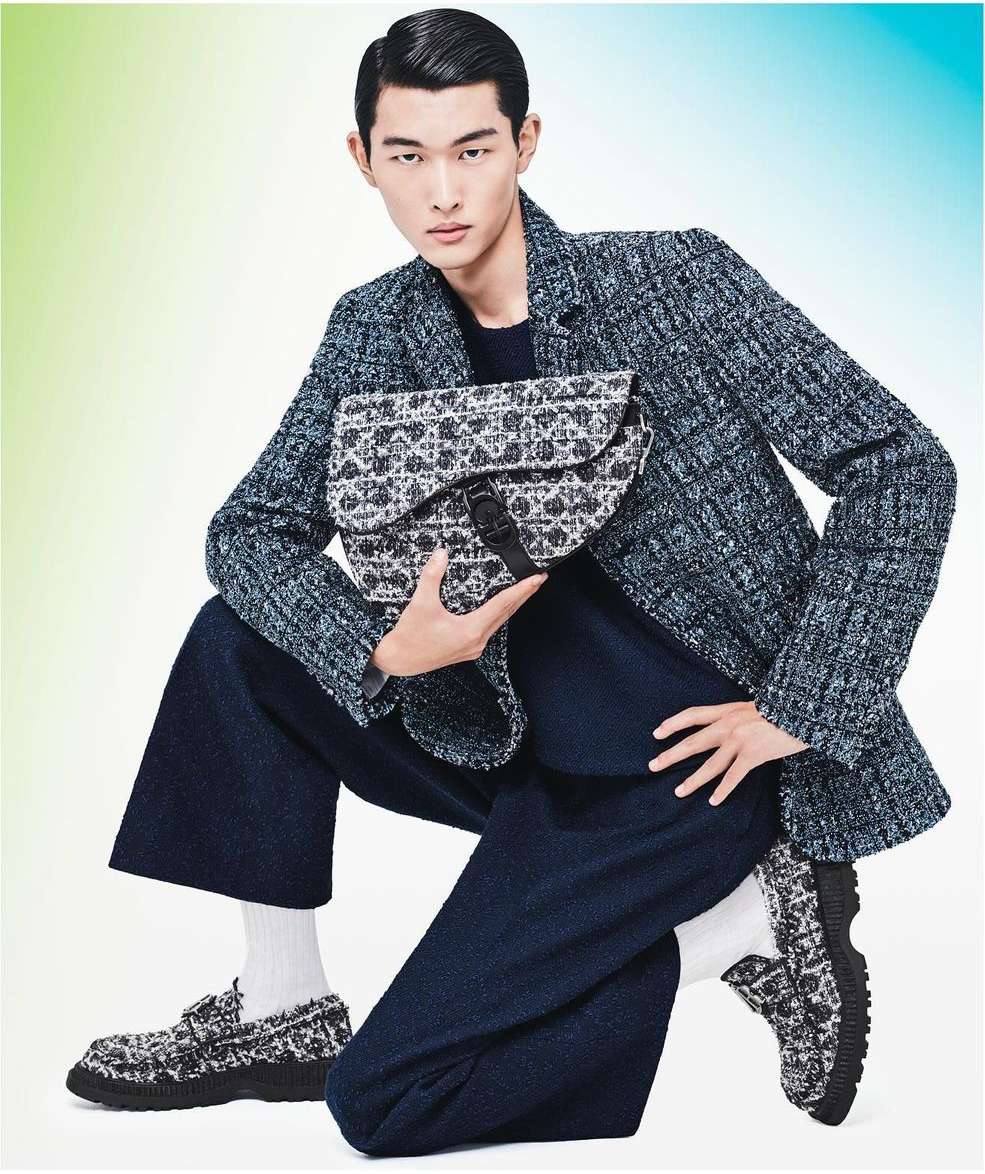 Asian man in fashion portrait wearing tweed jacket, navy culottes, and matching loafers, holding a printed saddle bag against a light background.