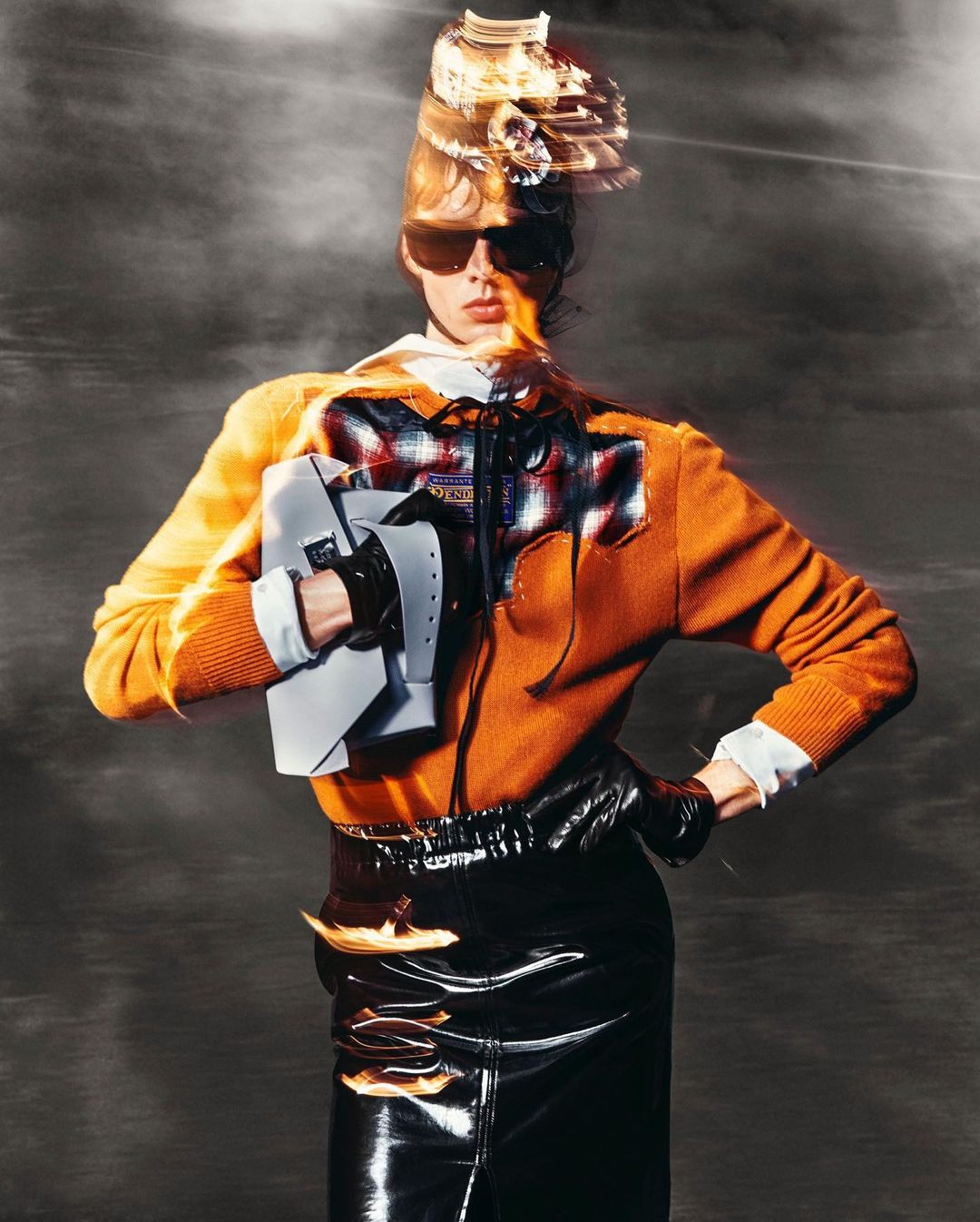 Fashionable woman posing in an orange top, black skirt, sunglasses, and unique hat against a hazy gray background in a studio shot.