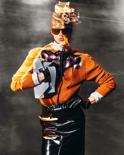 Fashionable woman posing in an orange top, black skirt, sunglasses, and unique hat against a hazy gray background in a studio shot.