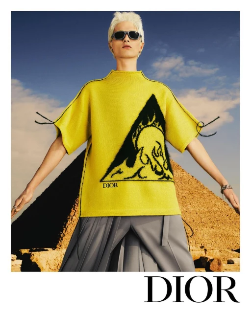 Male model in a yellow Dior top with a graphic print, grey pleated skirt, and sunglasses posing in front of a pyramid on a clear day.