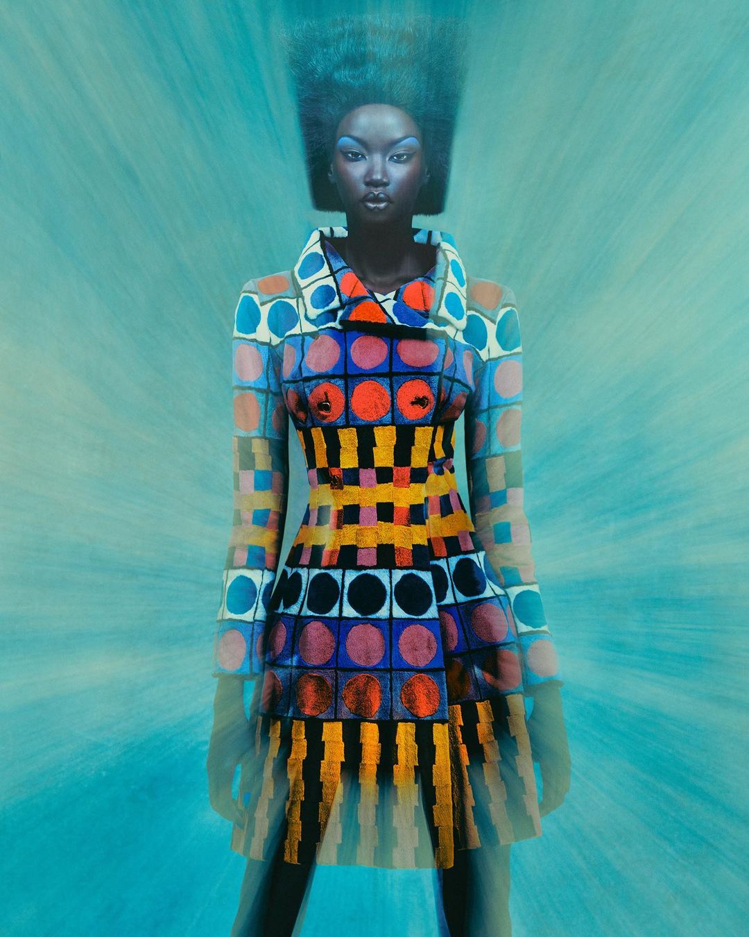 Black woman wearing a brightly patterned geometric coat and blue eye makeup against a teal textured background.