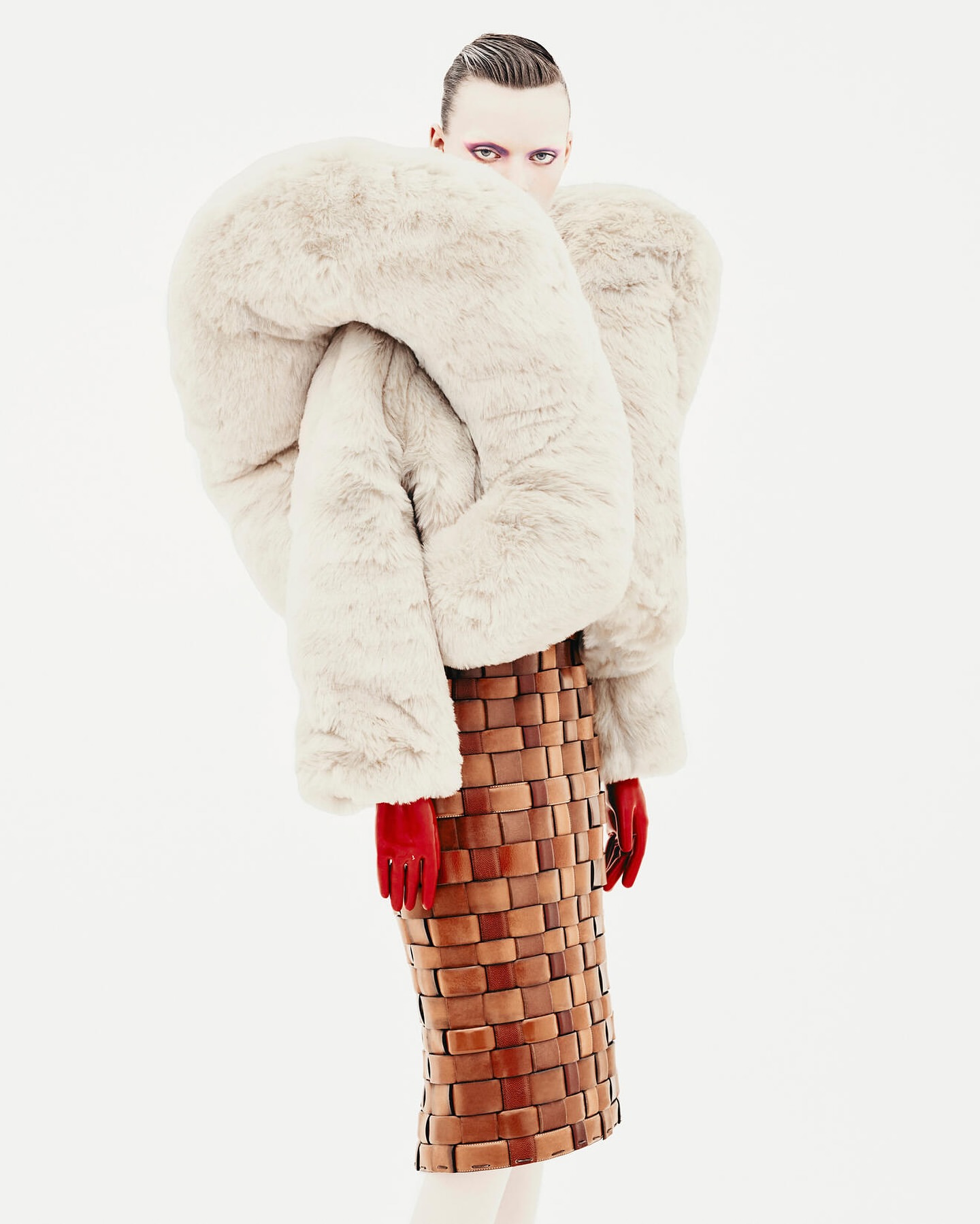 Avant-garde fashion portrait: Model with pale skin, dark eye makeup, wearing a sculptural fur coat and woven skirt in a stark white studio.