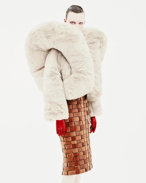 Avant-garde fashion portrait: Model with pale skin, dark eye makeup, wearing a sculptural fur coat and woven skirt in a stark white studio.
