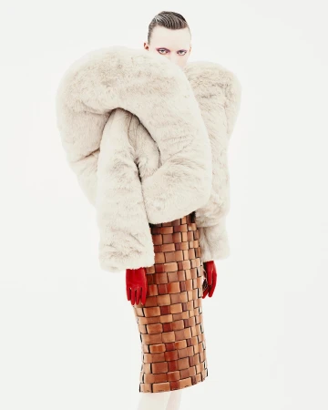 Avant-garde fashion portrait: Model with pale skin, dark eye makeup, wearing a sculptural fur coat and woven skirt in a stark white studio.