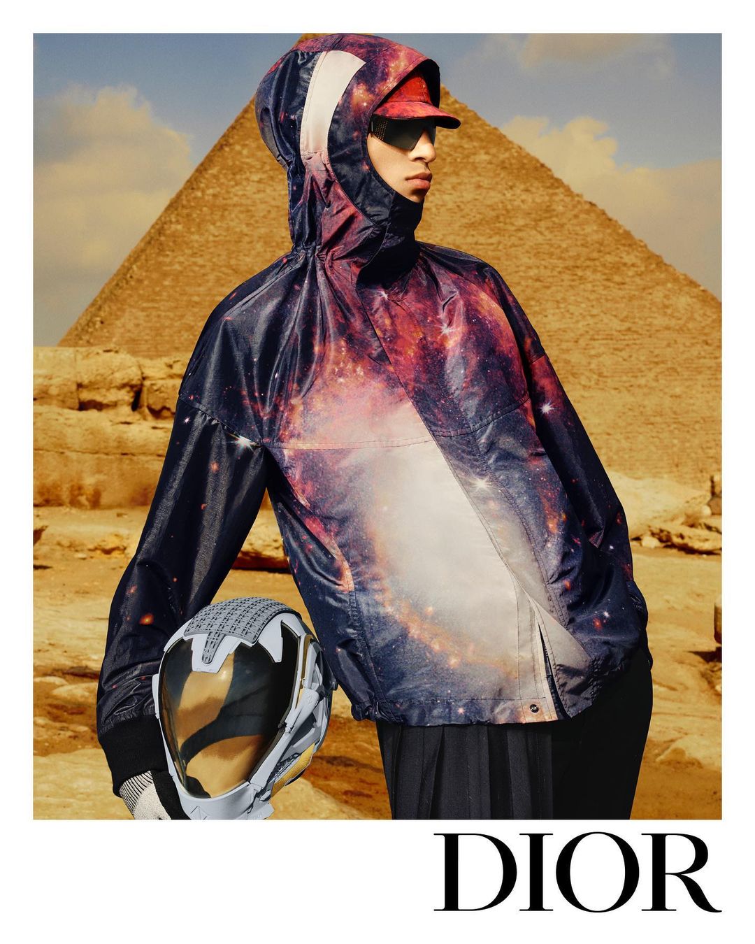 Fashion shot of a person wearing a hooded jacket with a galaxy print, holding a helmet, standing in front of a pyramid in Egypt.