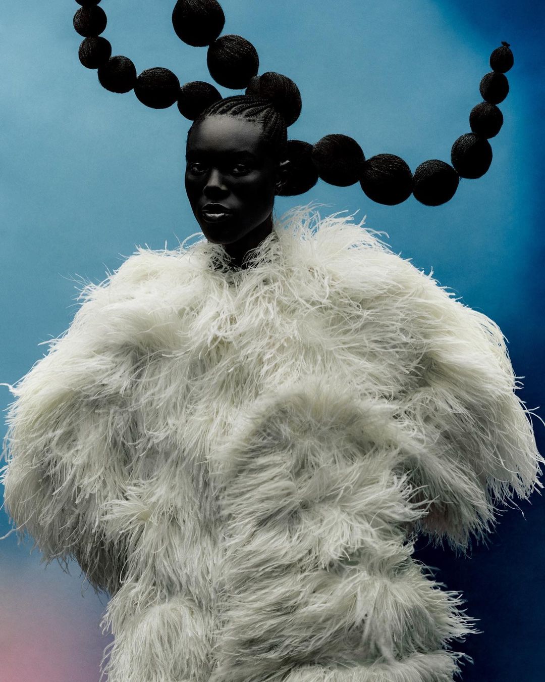 Fashion portrait of an adult female model with dark skin and braided hair, wearing a feathered coat with a blue gradient background.