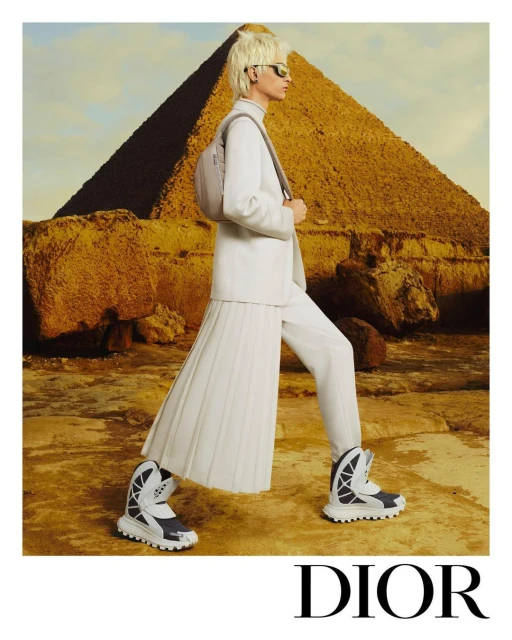 A model in white attire poses near the pyramid against a blue sky, showcasing Dior fashion with the Great Pyramid of Giza in the backdrop.