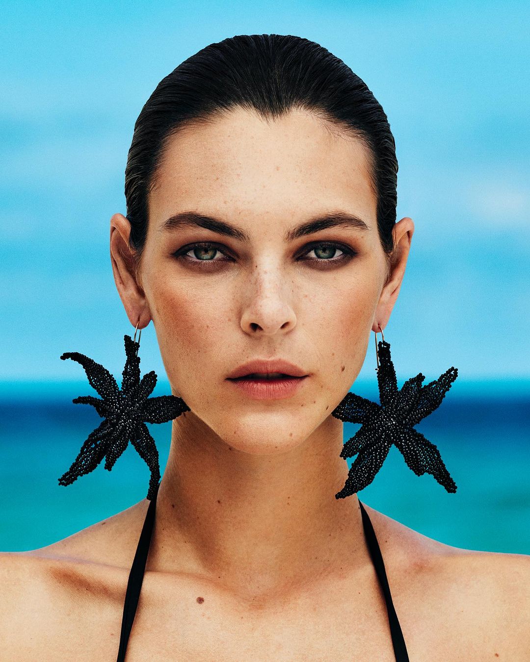Close-up portrait of a woman with slicked-back dark hair wearing large black star-shaped earrings against a bright blue ocean background