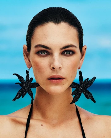 Close-up portrait of a woman with slicked-back dark hair wearing large black star-shaped earrings against a bright blue ocean background