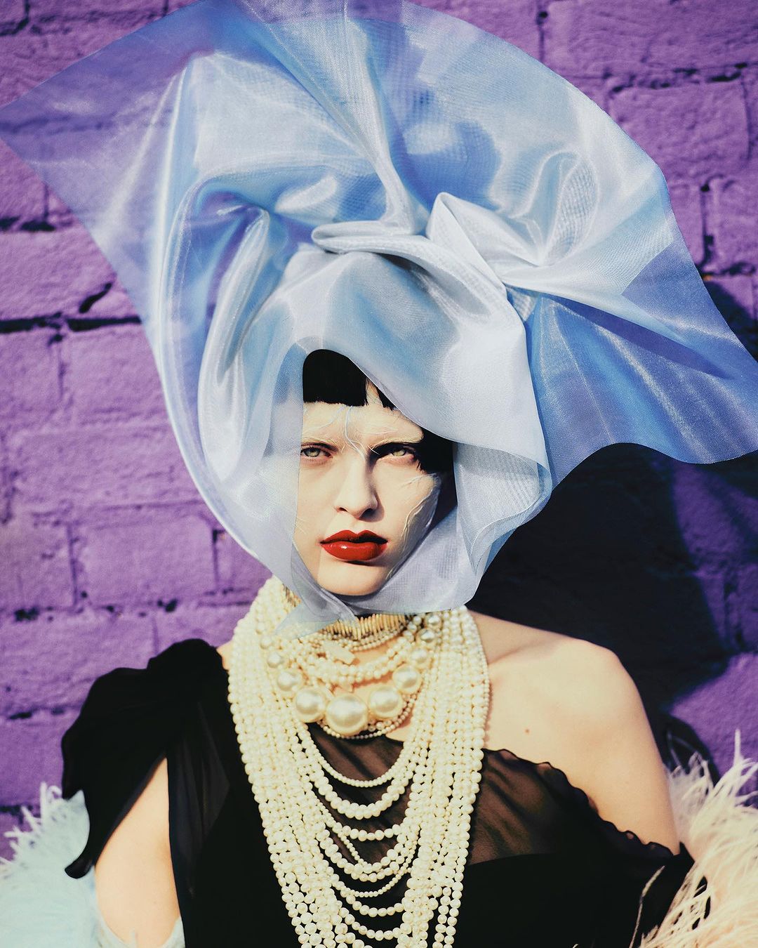 Fashion portrait of a woman wearing a light blue veil, pearl necklaces, and dark dress, standing against a purple brick wall background.