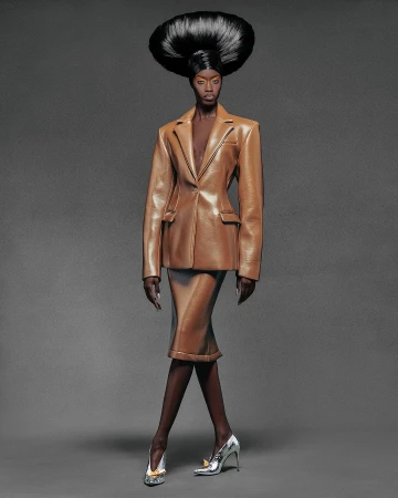 Model wearing a tan leather blazer and skirt with sculptural dark hair against a grey background