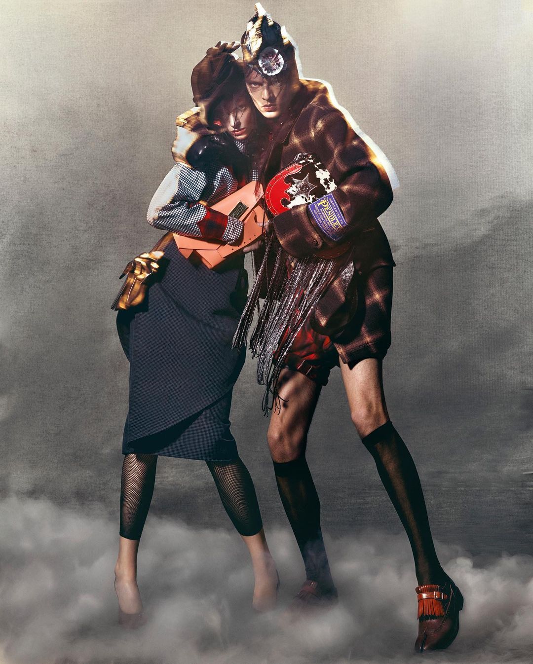 Two fashion models pose in an indoor studio setting with a textured background, showcasing unique clothing, handbags, and footwear.
