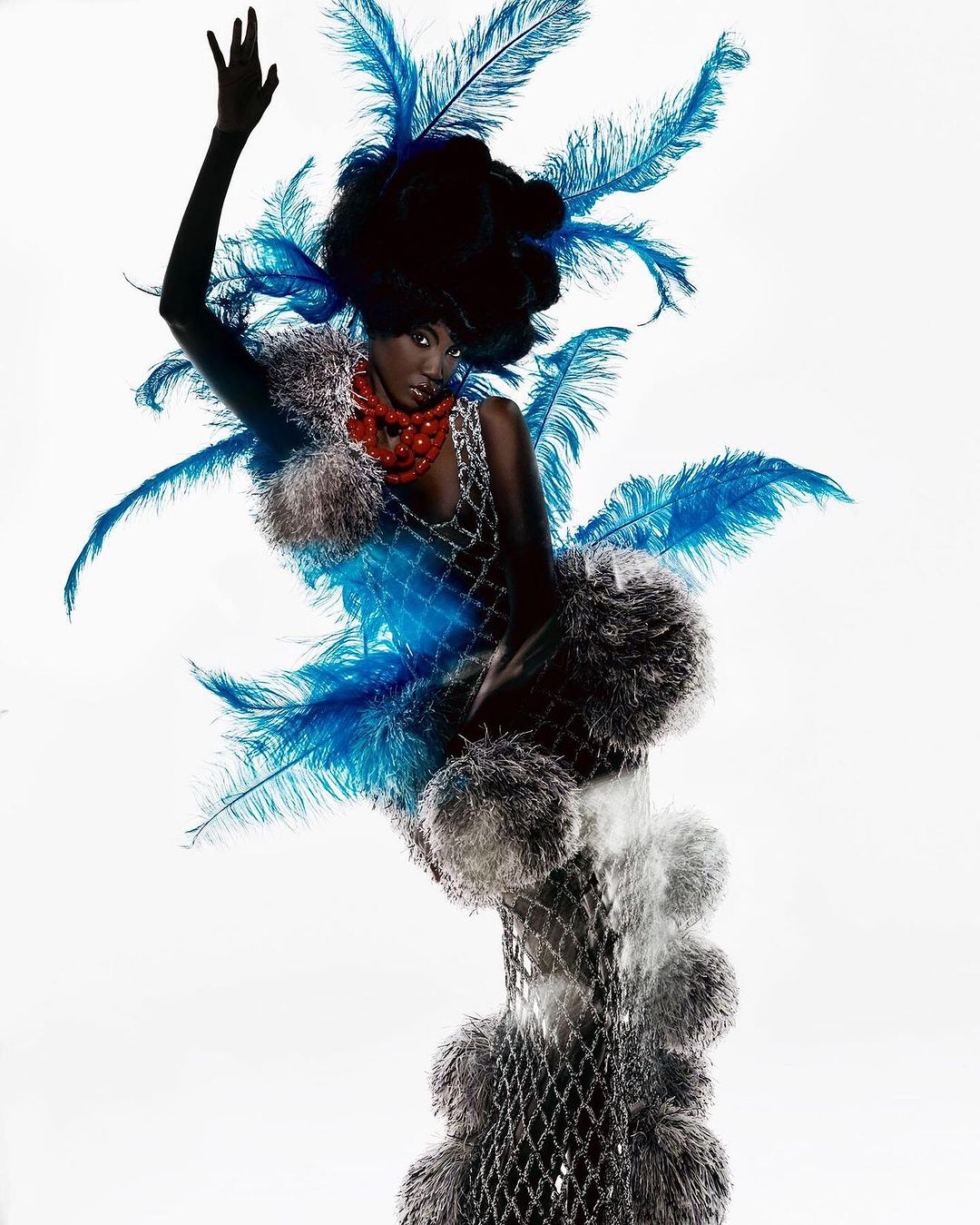 Fashionable woman in artistic portrait wearing feathers, fur, and statement jewelry against a plain white background in a studio shot.