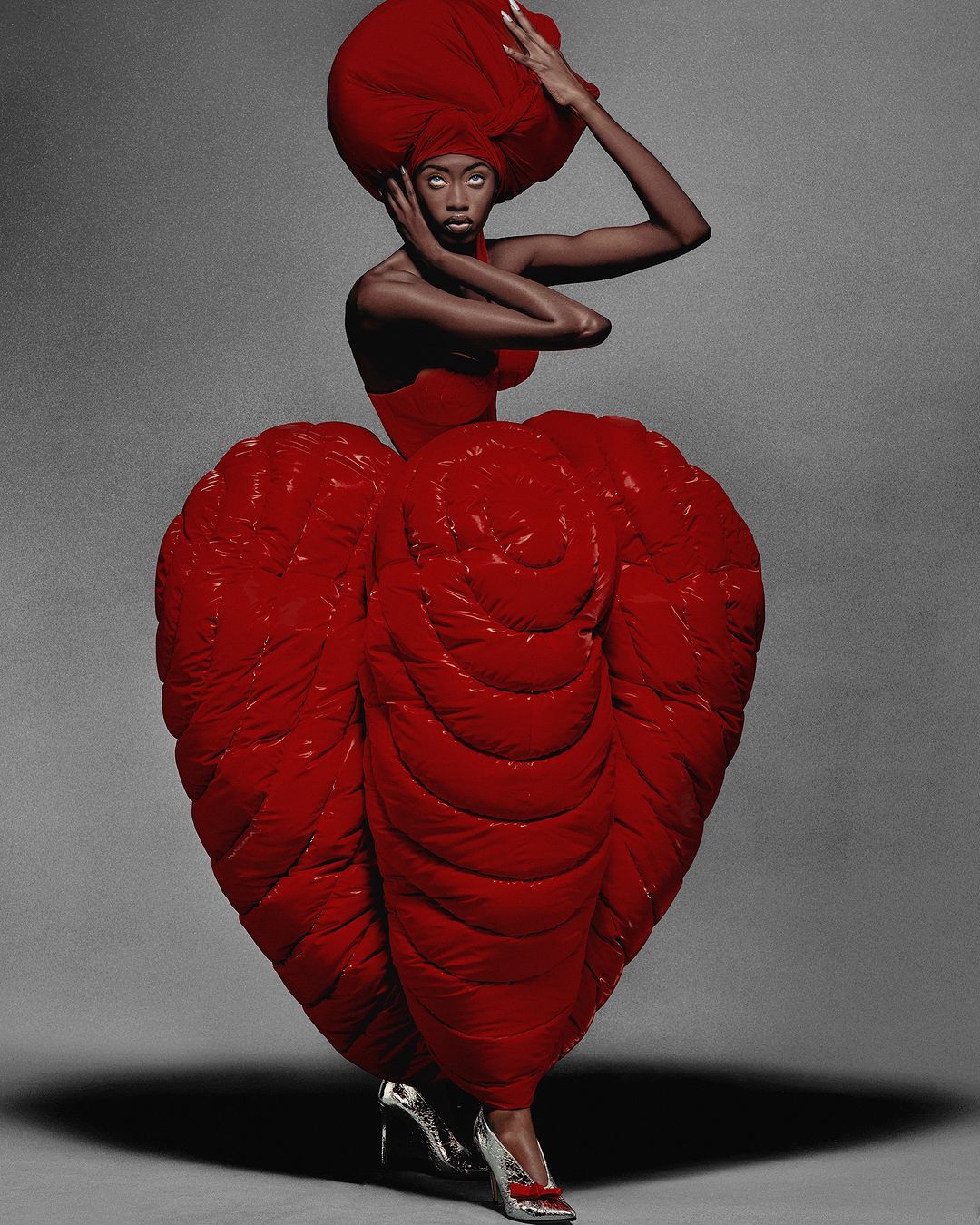 Editorial fashion portrait of an adult woman wearing a sculptural red gown and headpiece in a photo studio with soft, diffused lighting.