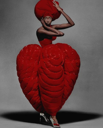Editorial fashion portrait of an adult woman wearing a sculptural red gown and headpiece in a photo studio with soft, diffused lighting.