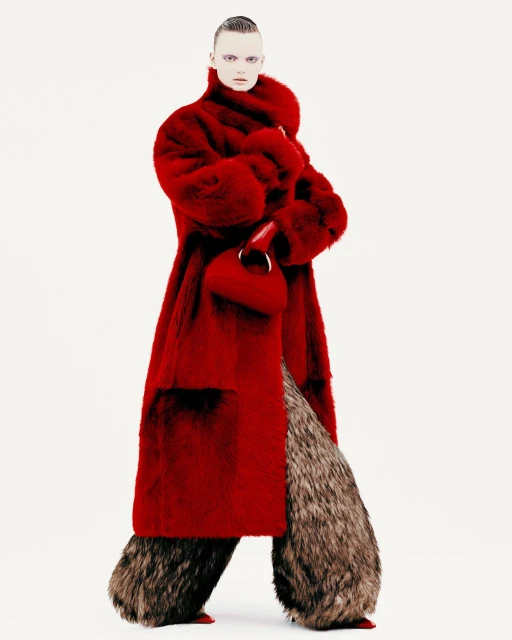 A fashion model stands in a light studio wearing a long red faux-fur coat and unusual fluffy brown pants with a red handbag.