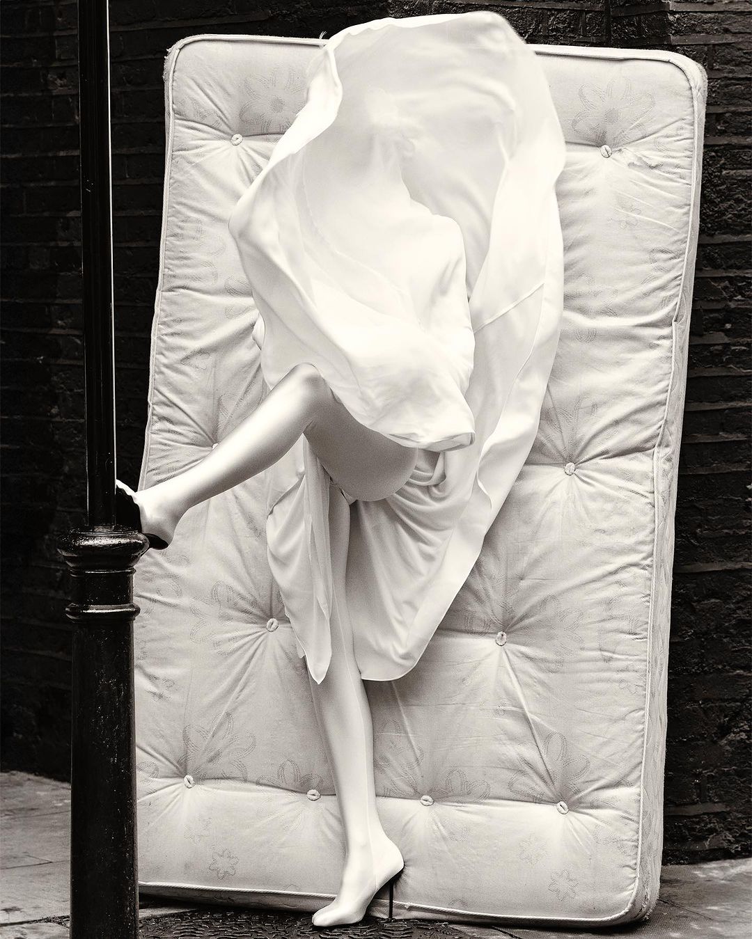 Surreal monochrome image: a woman's legs in high heels emerge from under a white sheet on a mattress, set against a brick wall.