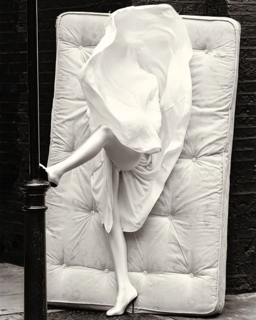 Surreal monochrome image: a woman's legs in high heels emerge from under a white sheet on a mattress, set against a brick wall.
