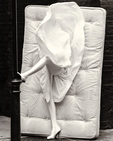 Surreal monochrome image: a woman's legs in high heels emerge from under a white sheet on a mattress, set against a brick wall.