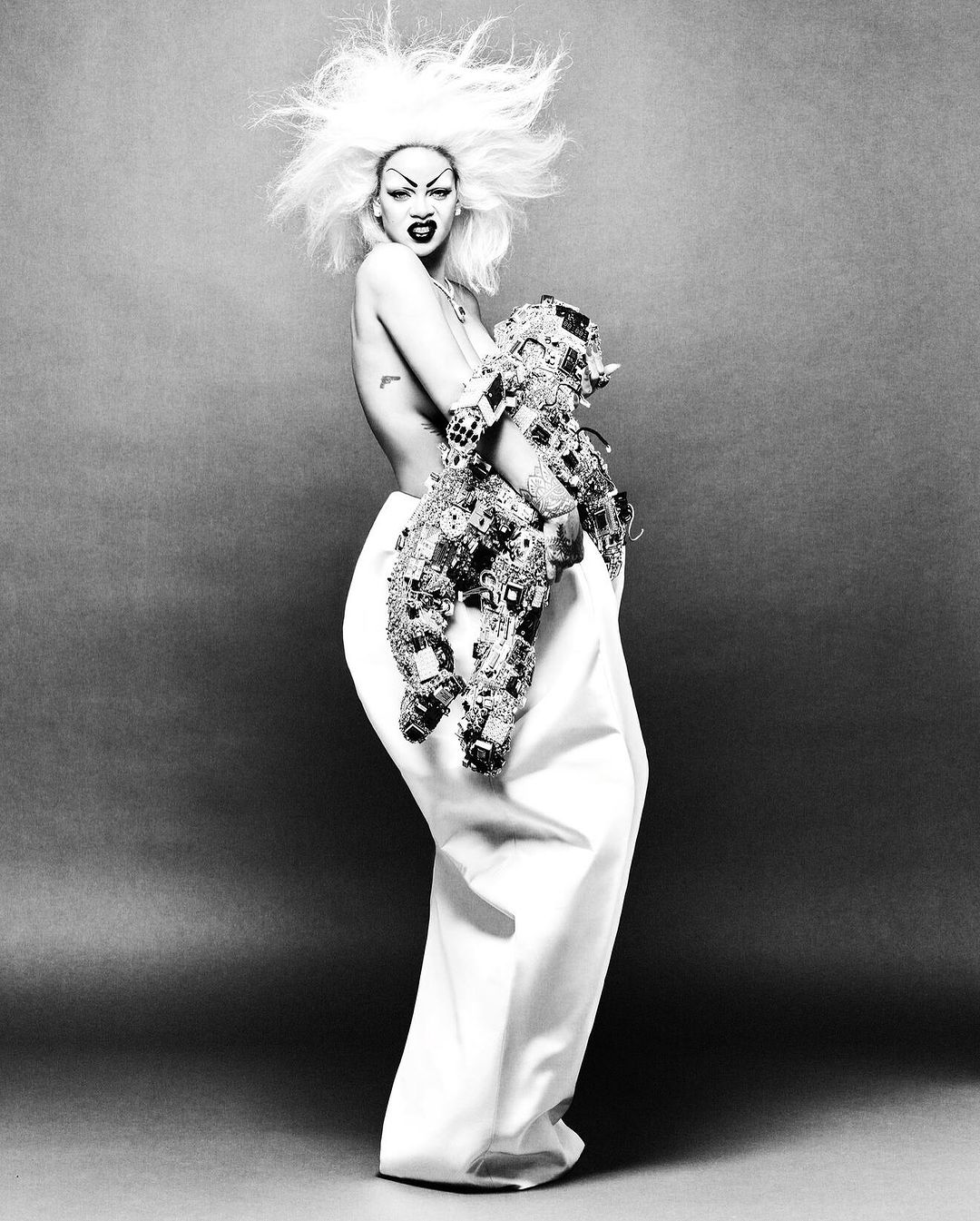 Striking black and white portrait of a person with elaborate hair and makeup, holding a decorative object and wearing a long skirt.