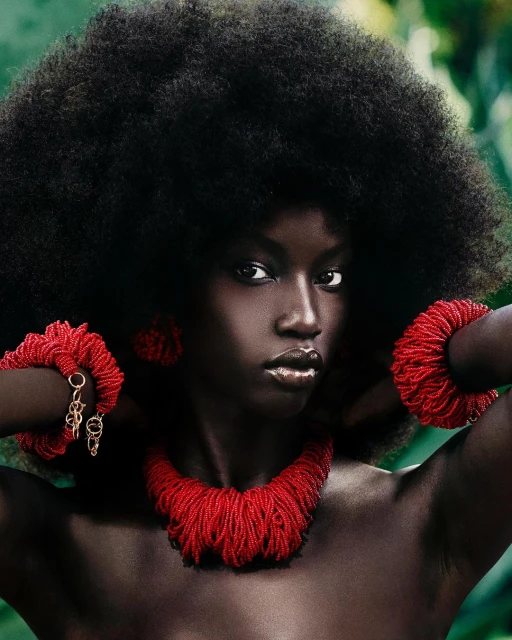 Portrait of a Black woman with a large Afro wearing a beaded red necklace and bracelets against a leafy green background in a beauty shot.