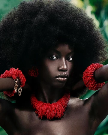 Portrait of a Black woman with a large Afro wearing a beaded red necklace and bracelets against a leafy green background in a beauty shot.