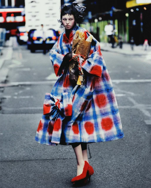 A model in an oversized, plaid coat and red heels walks down a city street holding wrapped baguettes in a fashion editorial shot.