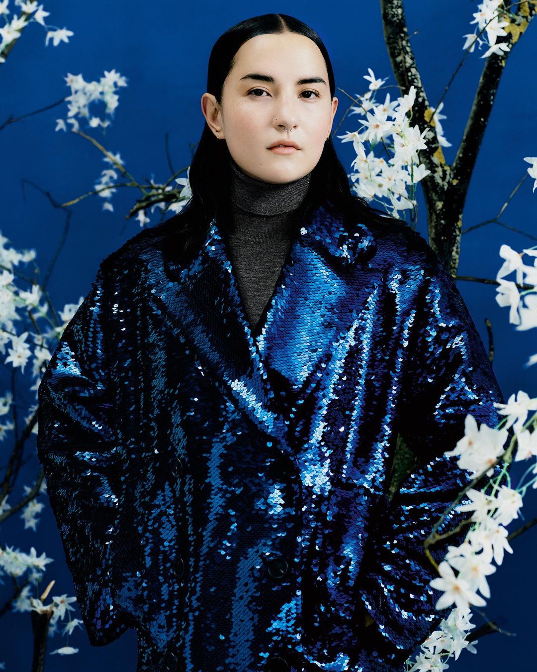 Portrait of a woman wearing a sequin coat against a blue background with flowering branches, posing in an indoor setting.